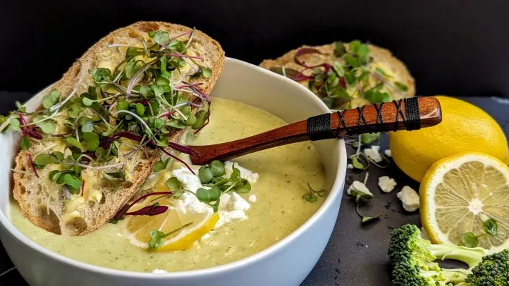 white bowl with green soup, topped with a crusty bread with butter and sprouts, in the background, some lemons and broccoli in front of a black background - Broccoli Feta Soup recipe